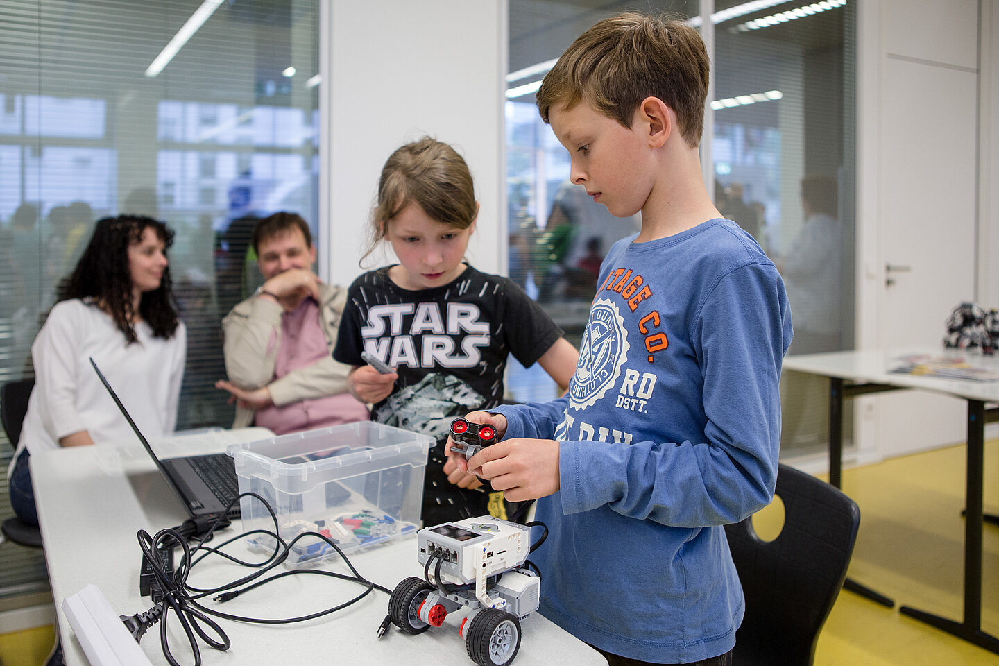 Im Vordergrund zwei Kinder, die einen Mini-Roboter bauen, im Hintergrund zwei Erwachsene im Gespräch