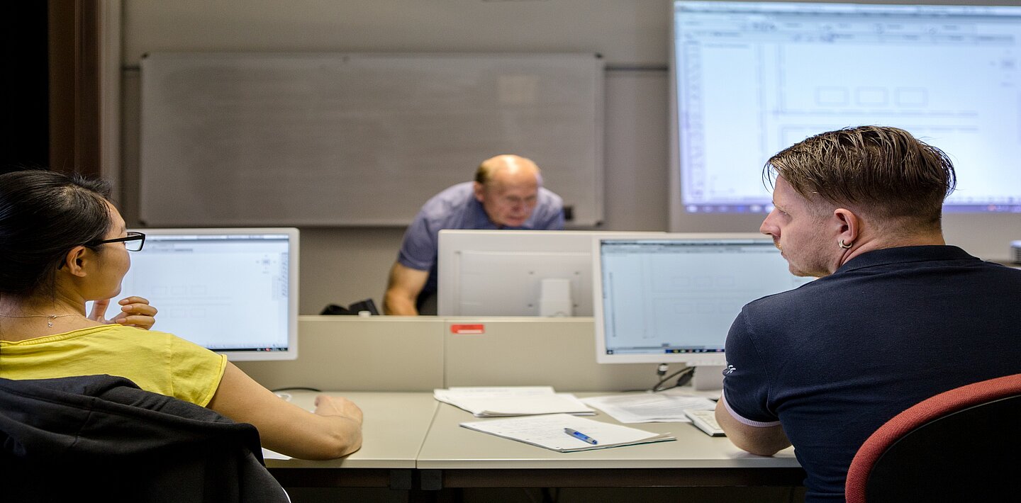 Während der AutoCAD-Schulung in der Volkshochschule (Foto: Robert Weinhold) Zwei nebeneinander sitzende Lehrgangsteilnehmer eines PC-Kurses tauschen sich aus, während der Kursleiter im Hintergrund über seinen Dozentenrechner gebeugt steht.