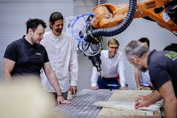 mehrere Männer stehen am Werktisch um einen Industrieroboter herum. Auf dem Tisch liegen Holzbretter.