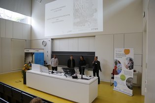 Vier Personen stehen am Rednerpult im Hörsaal. An die Wand wird eine Folie projiziert, und eine Frau spricht in ein Mikrofon