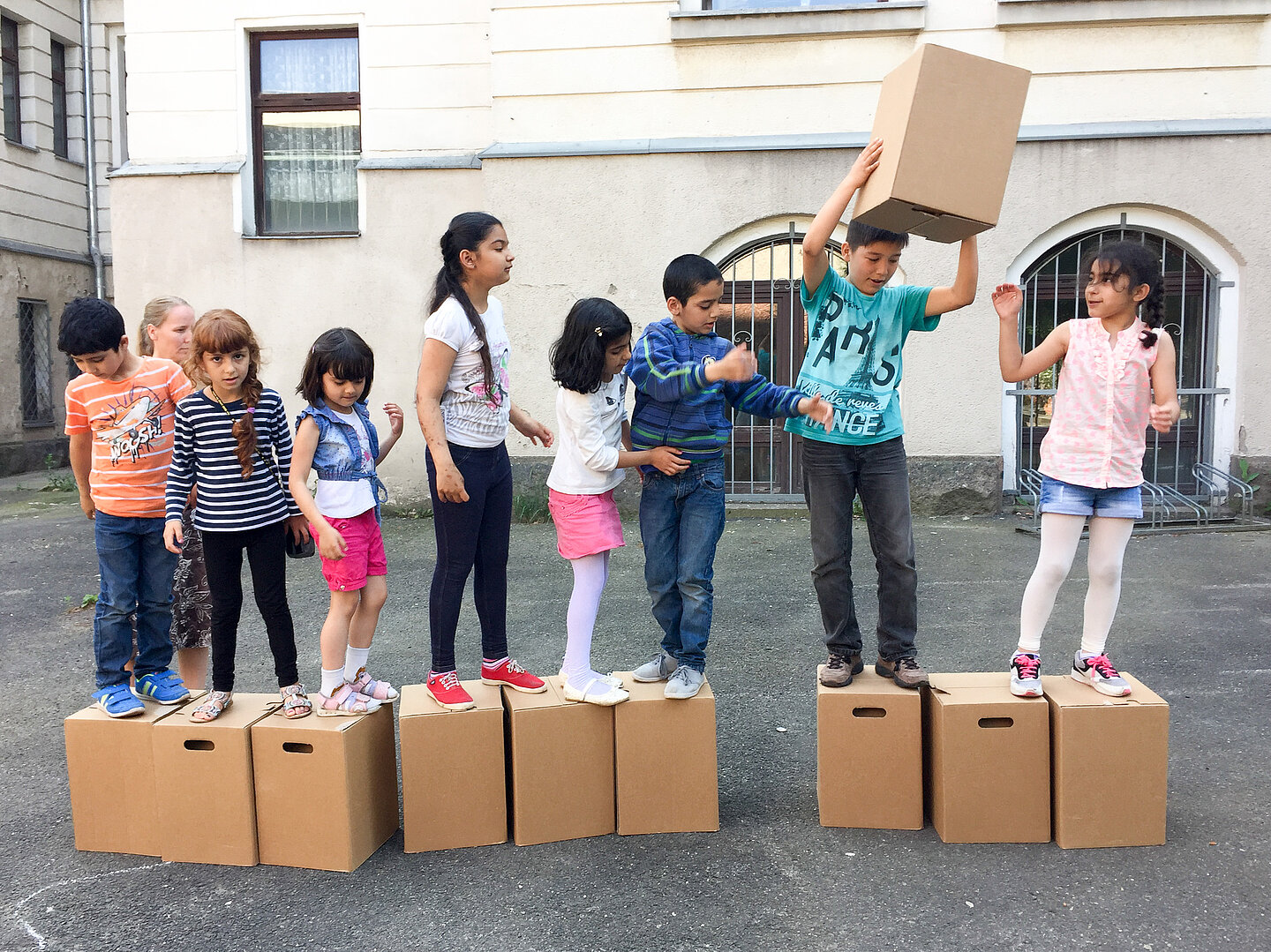 Gesundheitsförderung im Stadtteil Mehrere Kinder stehen in einer Reihe auf jeweils einem Pappkarton. Eines der Kinder gibt einen Pappkarton durch die Reihe nach vorne.