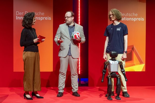 Moderatorin im Gespräch mit Prof. Crönertz, der einen Fußball hält, und Felix Loos mit humanoiden Roboter