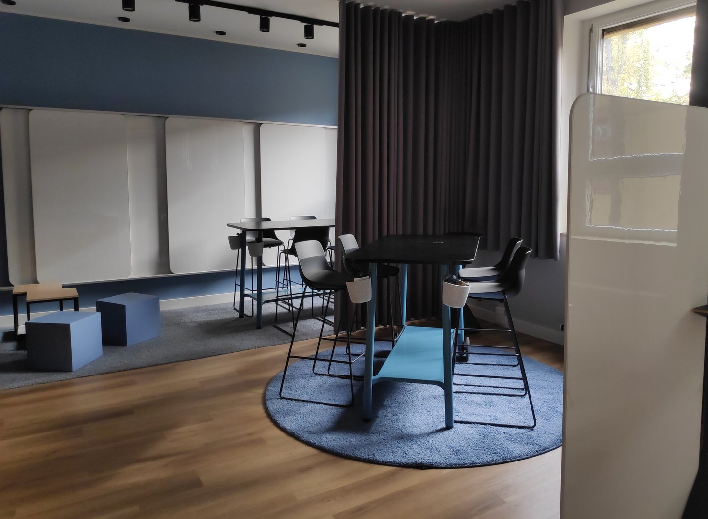 Die Lounge in einem entspannten Ambiente. Mehrere Sitzmöglichkeiten mit Hockern, Sitzwürfeln und Stühlen. Mehrere Whiteboards im Hintergrund.