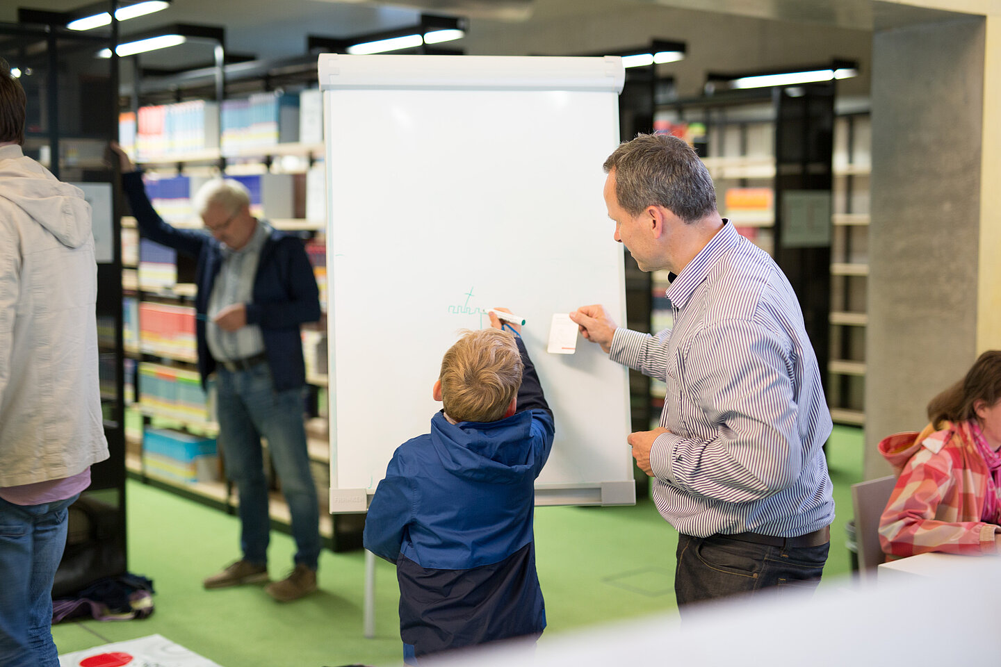Ein Kind und ein Erwachsender schreiben auf einem Flipchart, im Hintergrund Bücherregale und einzelne Menschen