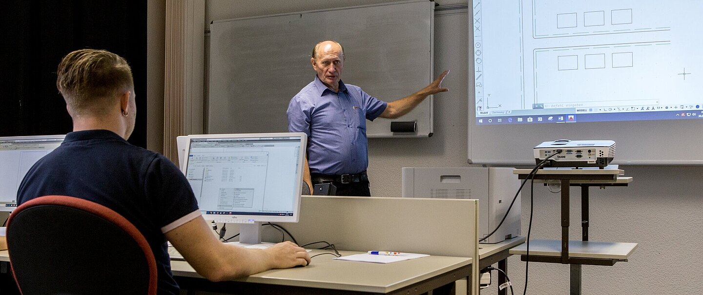 Während der AutoCAD-Schulung in der Volkshochschule (Foto: Robert Weinhold) Ein Dozent steht in einem Seminarraum vorn und zeigt auf eine Präsentation. Ein Lehrgangsteilnehmer sitzt mit dem Rücken zur Kamera vor einem Computer.