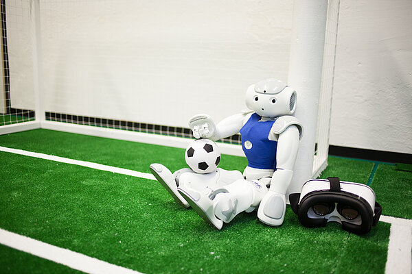 Nao-Roboter „Gargamel“ Nao-Roboter „Gargamel“ sitzt an einem Tor mit Fußball und VR-Brille daneben.