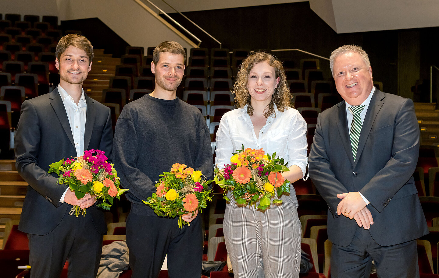 Preisverleihung Zwei Männer und eine Frau je mit Blumenstrauß in der Hand sowie ein weiterer Mann stehen auf der Bühne im großen Saal des Gewandhauses
