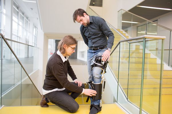 Max hat Exoskelett Prototyp am linken Bein, Felix justiert daran/bei Verlinkung führt Link zur Projektwebsite